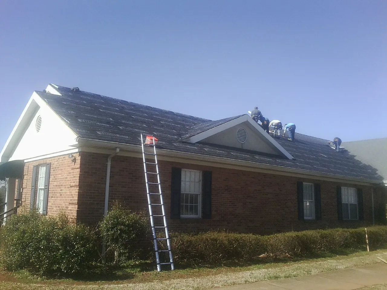 Crew tearing shingles off a brick ranch roof with a ladder set — Metal Roof Repair work by our crew in Bernard