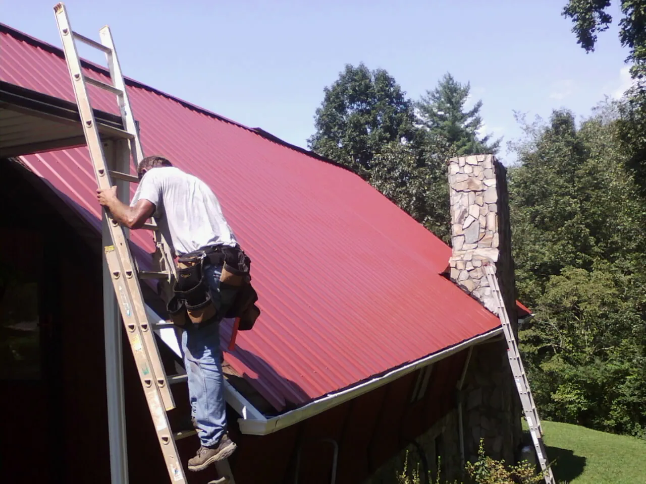 Roofer on a ladder beside a red metal roof and stone chimney — Soffit & Fascia Repair by Roofing Brothers in Bernard